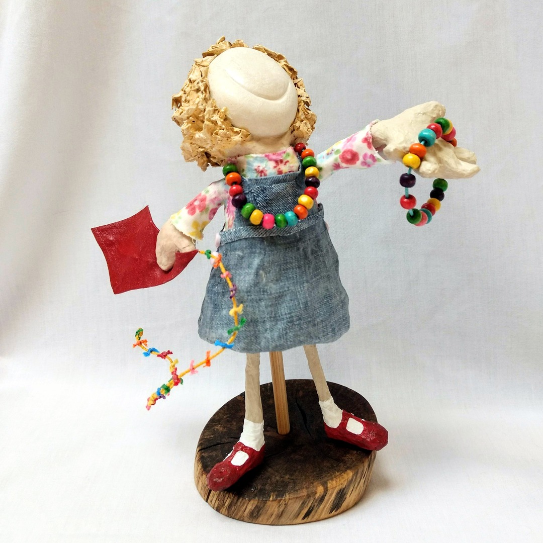 mixed media doll of a girl with a kite. she is offering you a necklace matching her own.